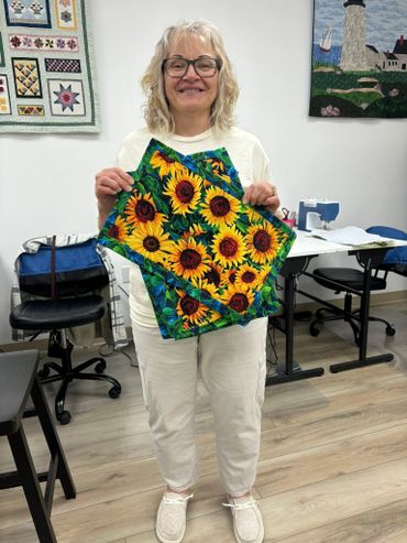 Smiling woman proudly holds a sunflower-patterned quilt piece in a sewing room.