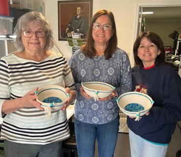Three women proudly holding handcrafted bowls with blue interiors indoors.