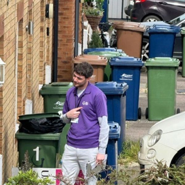 Man in purple shirt stands near colorful trash bins on a residential street.