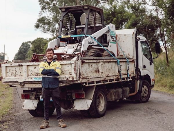 Man standing confidently in front of a work truck carrying a Bobcat loader on a rural road.