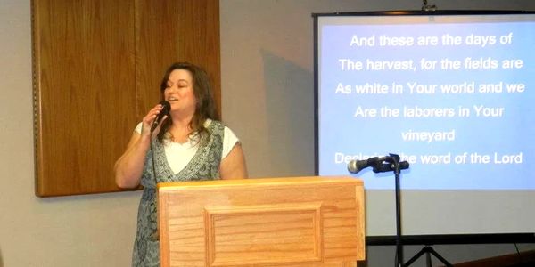 Woman singing at a podium with religious lyrics projected.