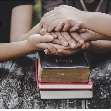 Hands joined in prayer over a Holy Bible and two other books.