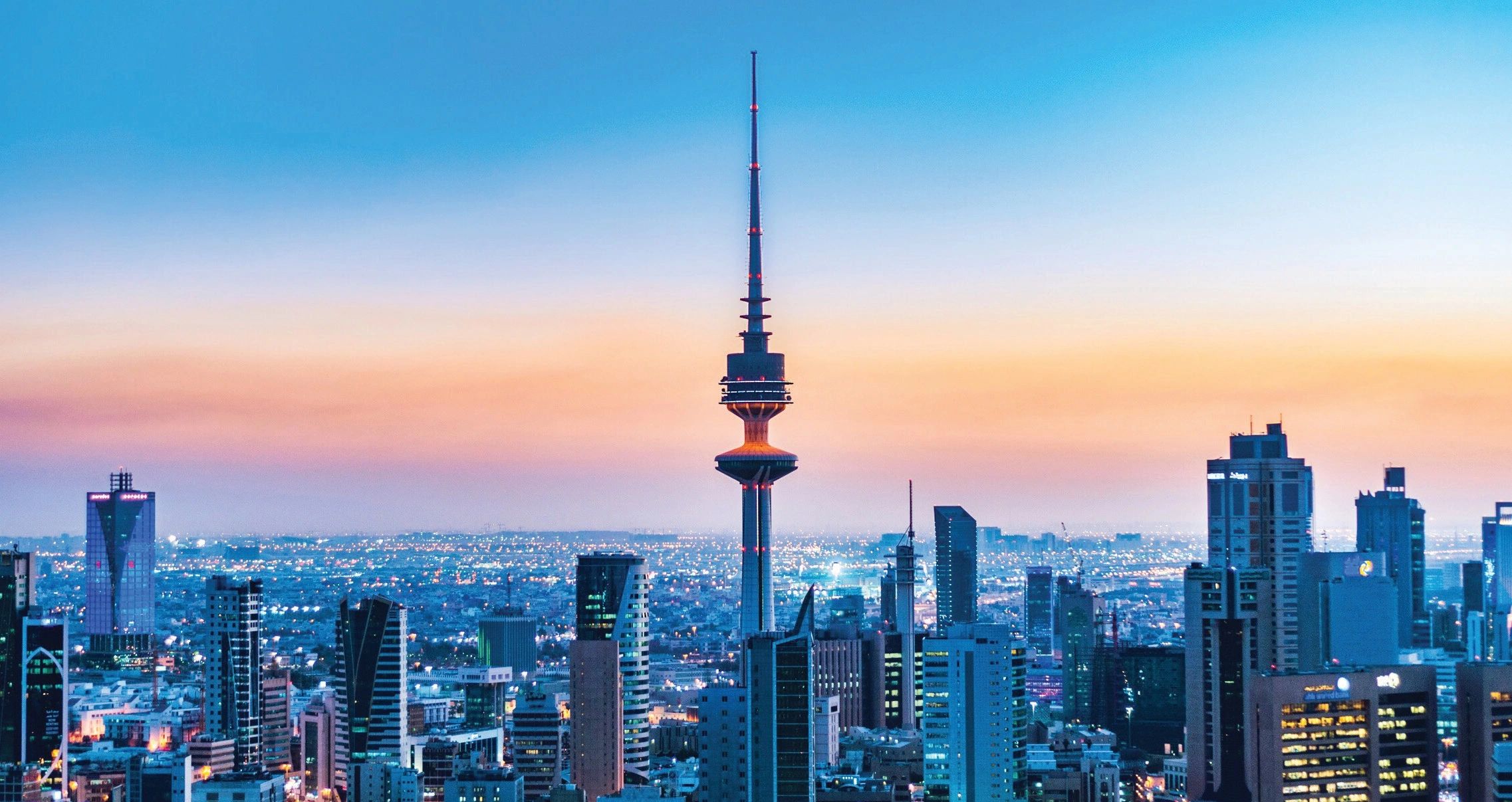 Skyline of a modern city with a tall communication tower during sunset.