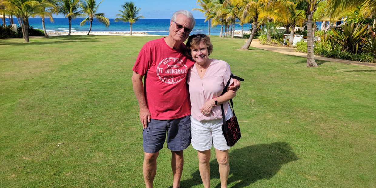 Smiling couple posing on a grassy lawn by the ocean and palm trees.