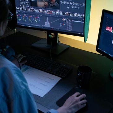 Person editing video color grading on dual monitors in a dark room.