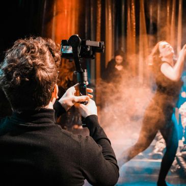 A person filming a dancer in a smoky, dimly lit stage environment.