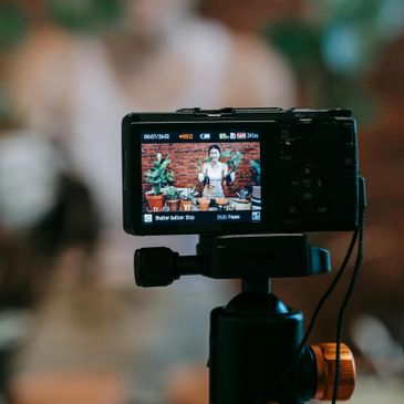 Camera screen shows a woman holding plants, recording a video indoors.