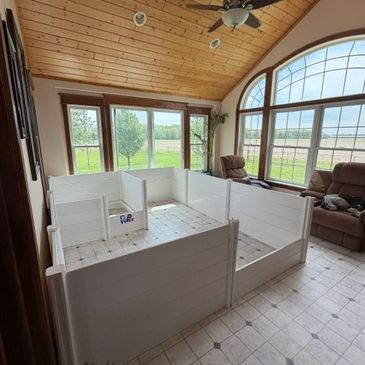 Indoor white wooden playpen in a sunlit room with large windows and recliners.