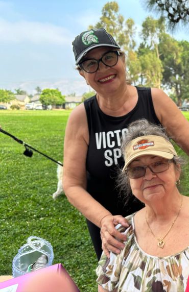 Two smiling older women outdoors, one standing and one seated, enjoying a sunny day in the park.