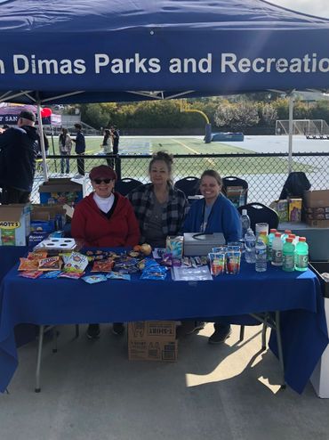 Three women sit under a canopy at a snack and drink table during an outdoor event.