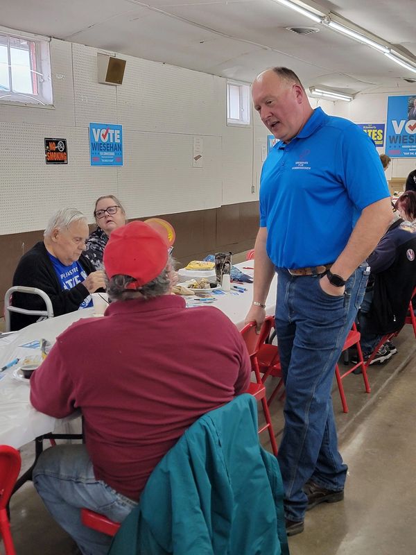 A man in blue talks to seated people at a community event with voting posters.