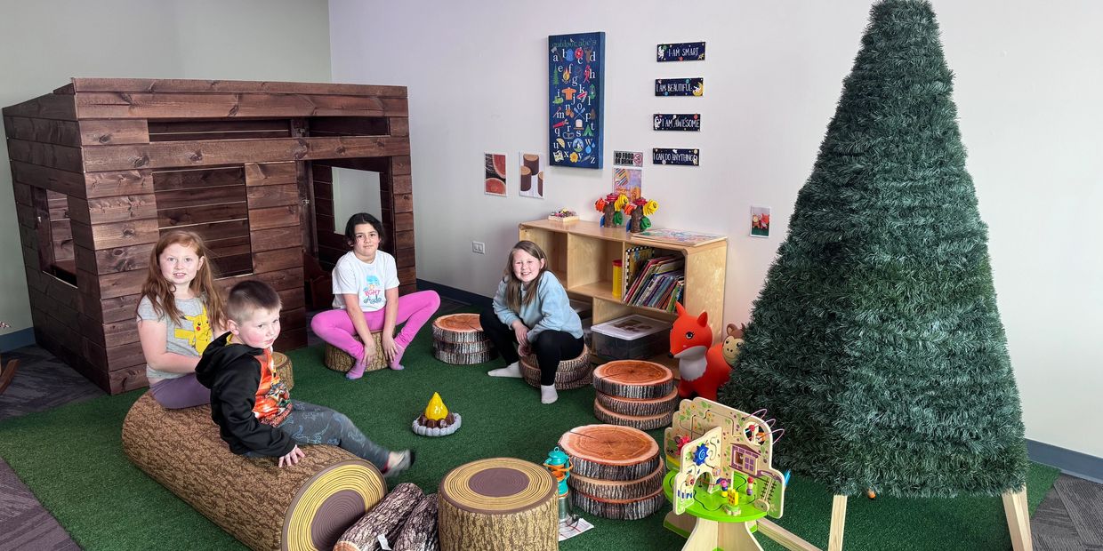 Children sitting on log-themed seats in a cozy indoor play area with a wooden playhouse and artificial tree.