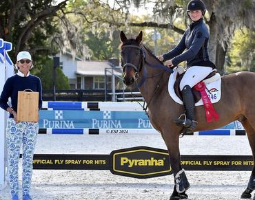 Equestrian rider on horse with a red ribbon, standing next to an award presenter outdoors.