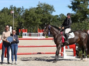 Equestrian rider on horse with two women holding a trophy beside a jump.