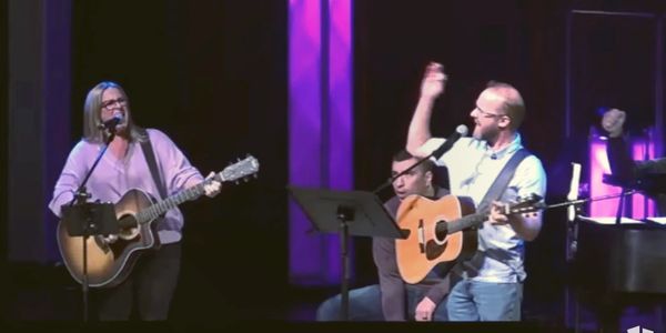 Two musicians passionately playing guitars and singing on stage with purple lighting.