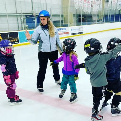 Ice skating instructor teaches young children on rink, wearing helmets and protective gear.