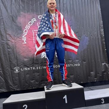 Athlete stands on first place podium, draped in American flag.