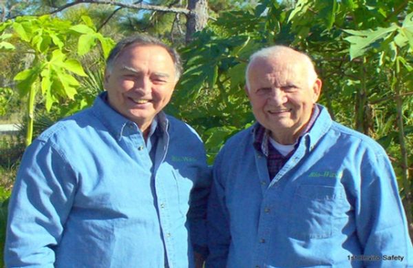 Two older men smiling outdoors in matching blue shirts.