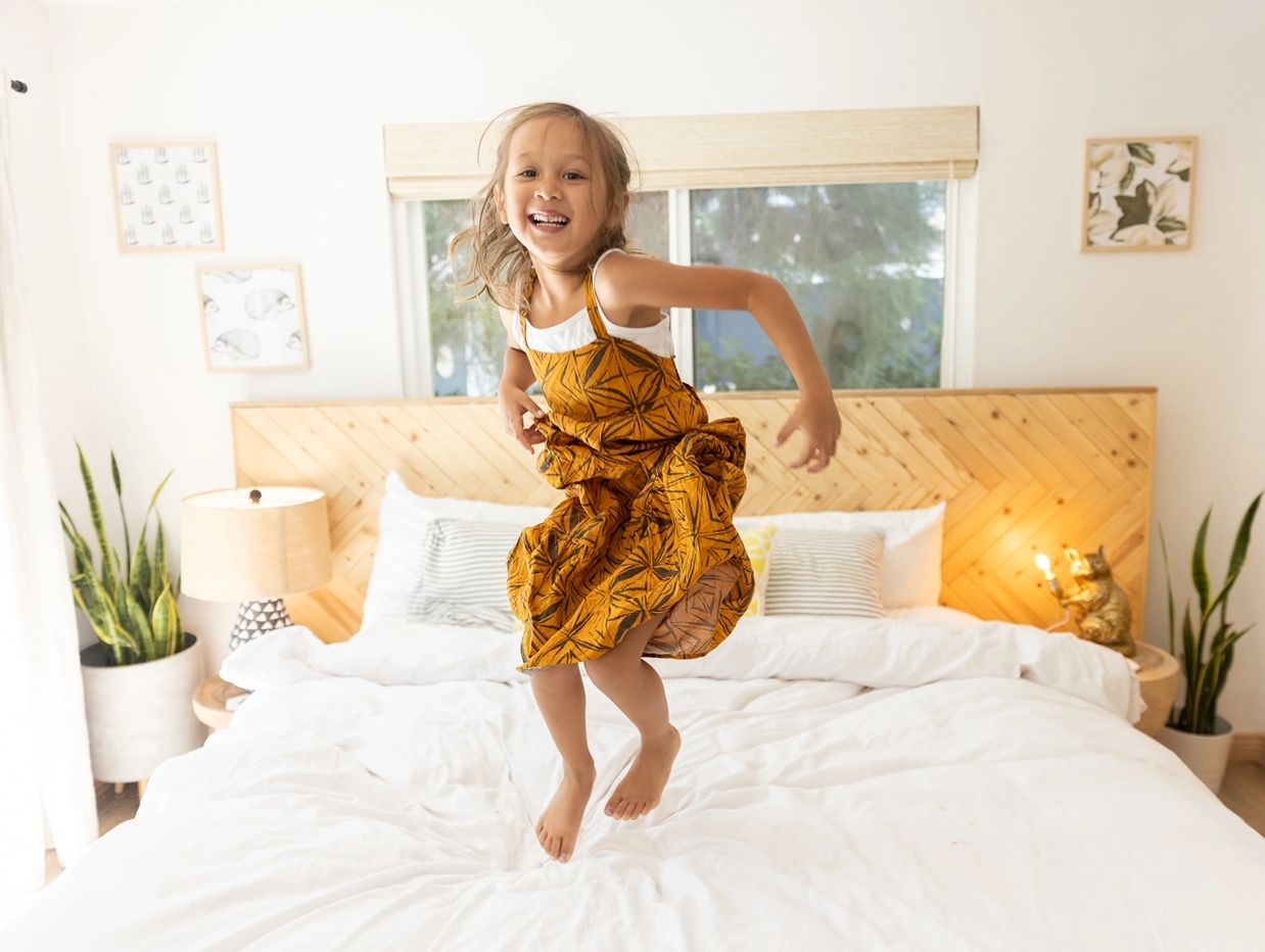 young child in sustainably designed golden brown dress jumping joyfully on a white cotton bed