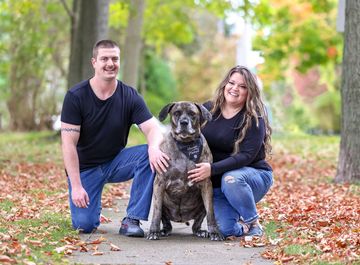 A happy couple kneels on a sidewalk with their large dog in an autumn park.