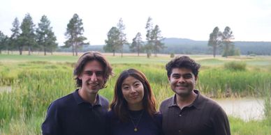 Three friends smiling outdoors with a scenic nature background of trees and grass.
