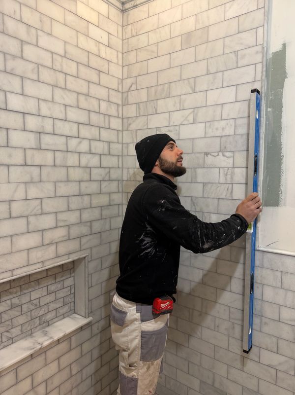 A man wearing a black beanie and sweater uses a level tool in a tiled bathroom.