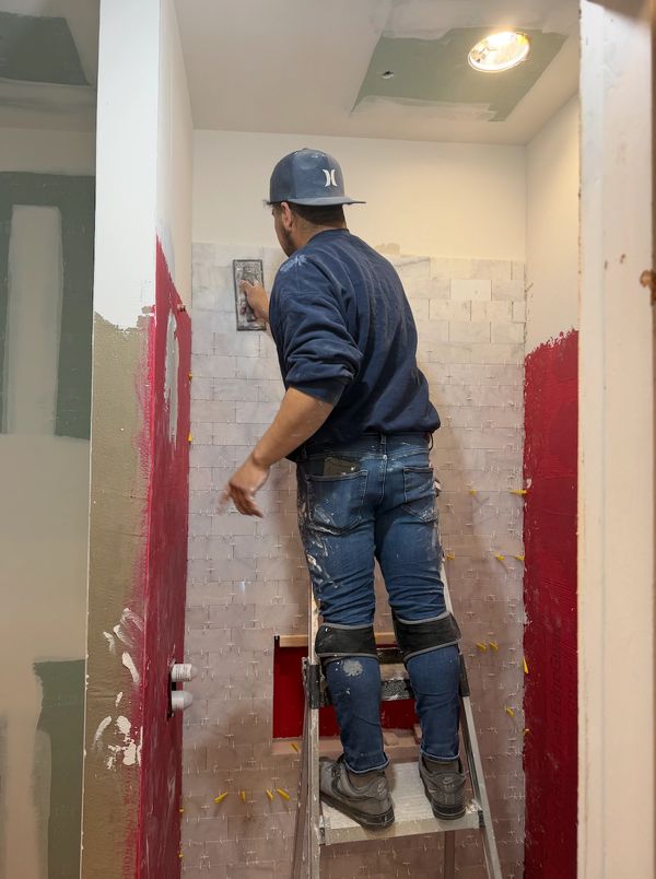 Man tiling a small bathroom wall while standing on a ladder.