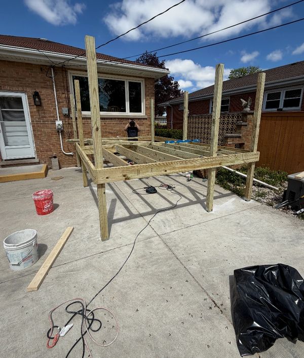 Wooden deck frame being constructed on a concrete patio beside a brick house.