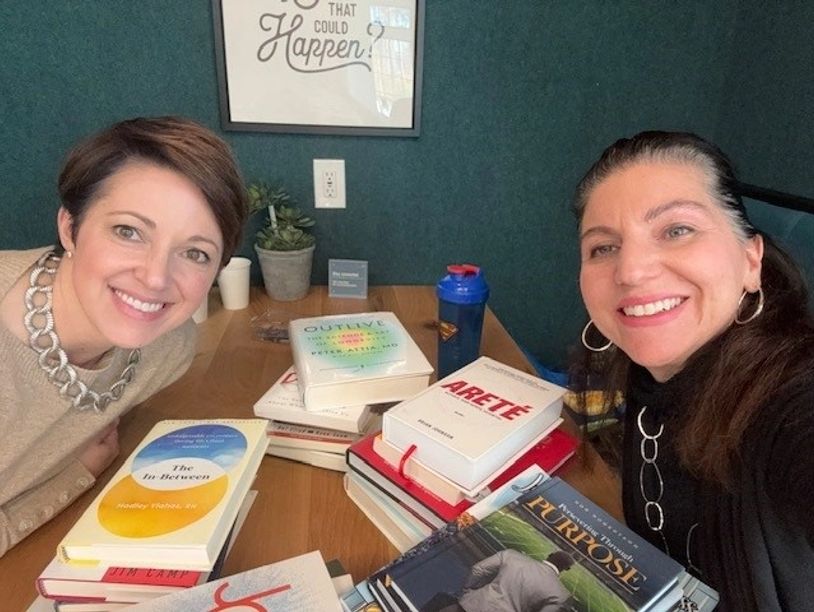 Two smiling women at a table with stacks of books in a cozy reading room.