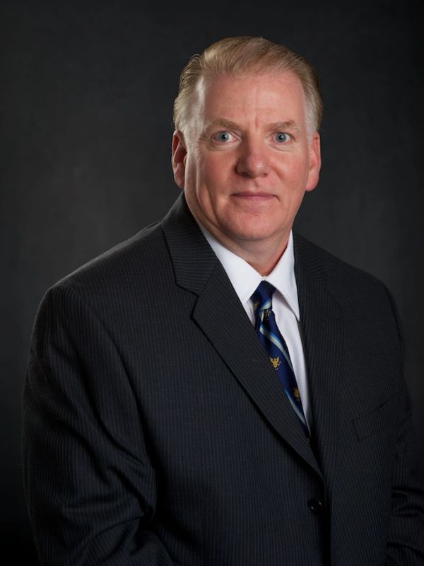 Professional portrait of a man in a dark suit and blue tie against a black background.