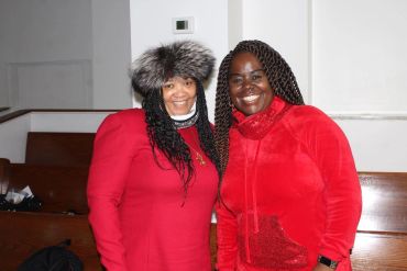 Two women smiling, dressed in red, one wearing a furry hat indoors.
