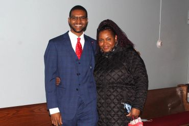 A man in a blue suit and red tie poses with a woman in a black quilted jacket indoors.