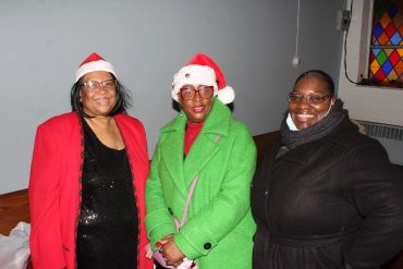 Three women smiling indoors, two wearing Santa hats and festive coats.