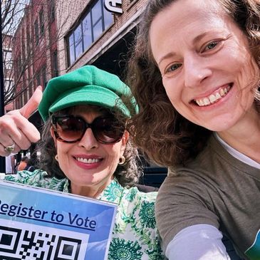 Two women smiling, one holding a 'Register to Vote' sign with a QR code outdoors.