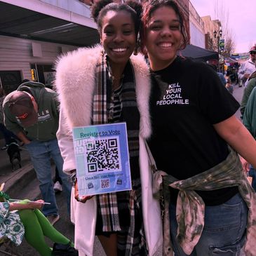Two smiling women, one holding a voter registration QR code poster.