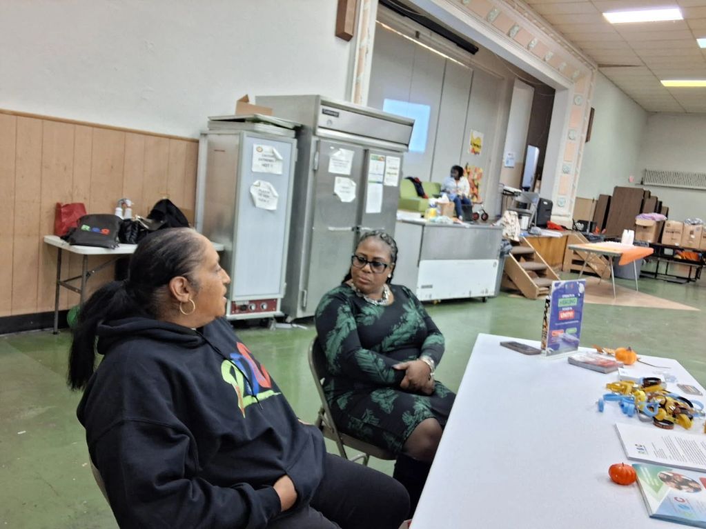 Two women sitting and talking in a community hall with tables and supplies.
