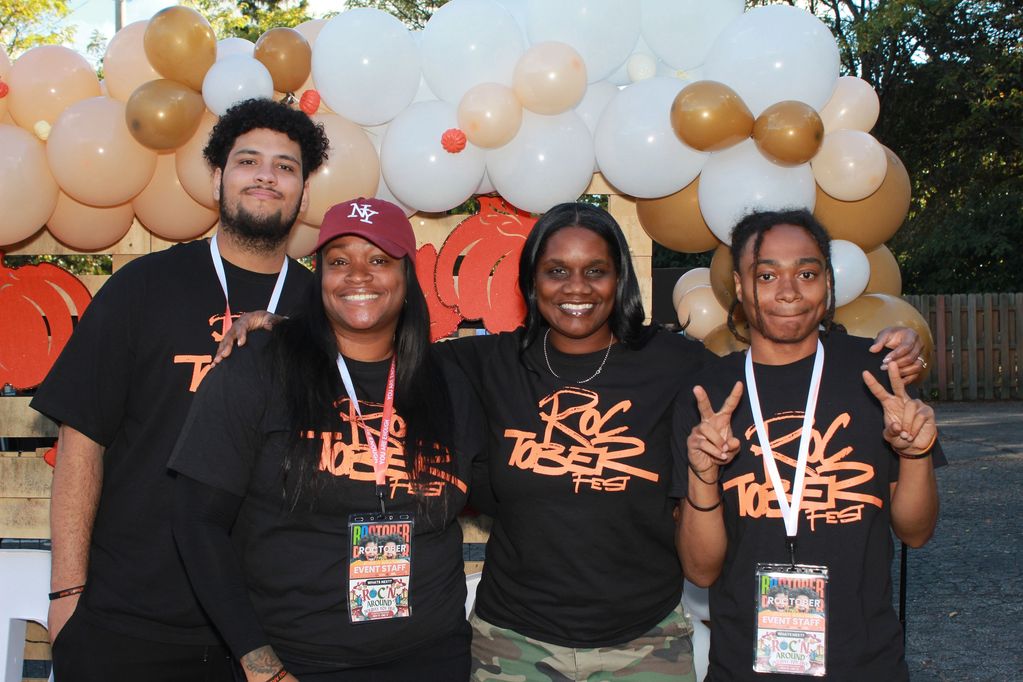 Four smiling event staff in matching Roc Together Fest shirts with festive balloons background.