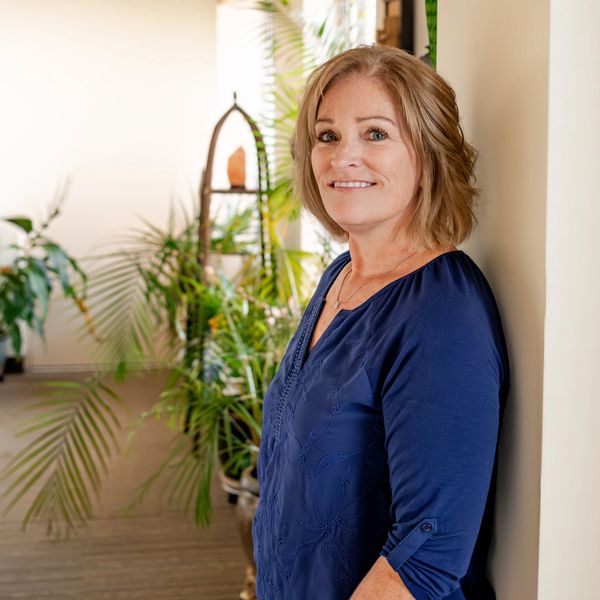 Smiling woman in a blue shirt leaning against a wall with plants in the background.