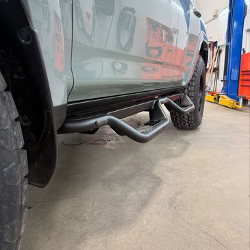Black side step bars installed on a white off-road vehicle in a garage.