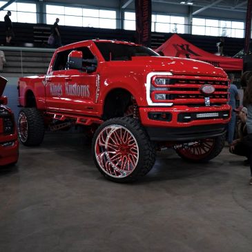Bright red lifted custom truck with large chrome wheels at an indoor auto show.