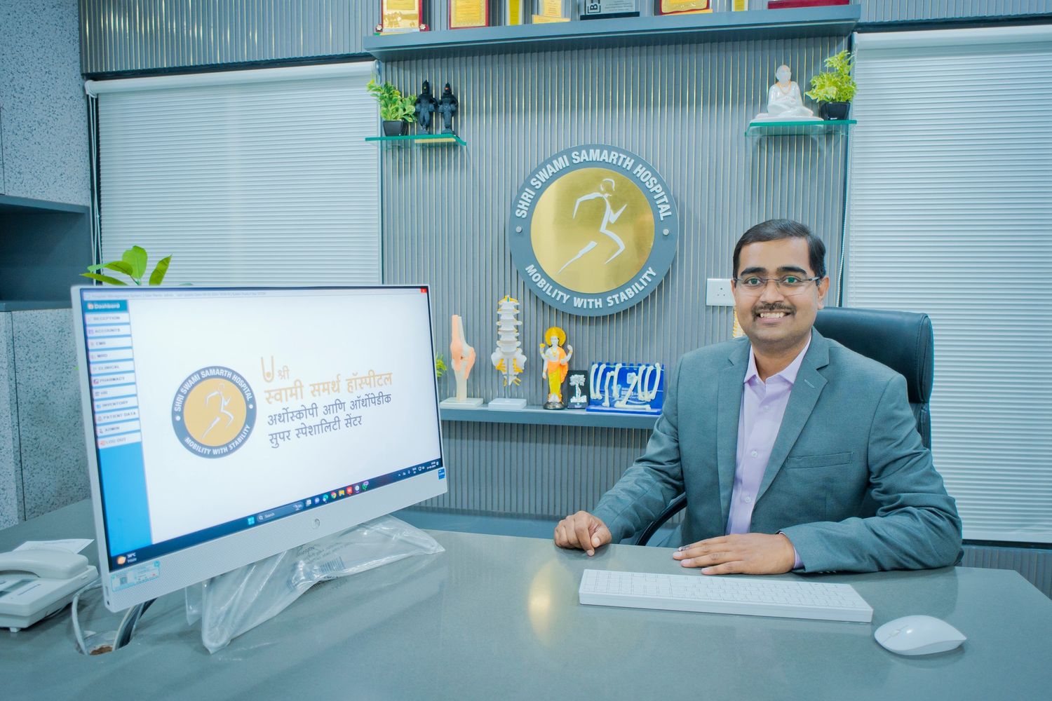 A smiling man in a suit seated at a hospital desk with a computer.
