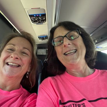 Two women smiling in pink shirts inside a vehicle.