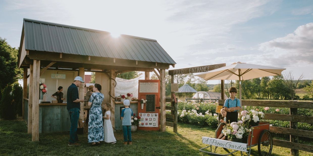 People enjoying a sunny day at a rustic garden entrance with flowers and a small stand.