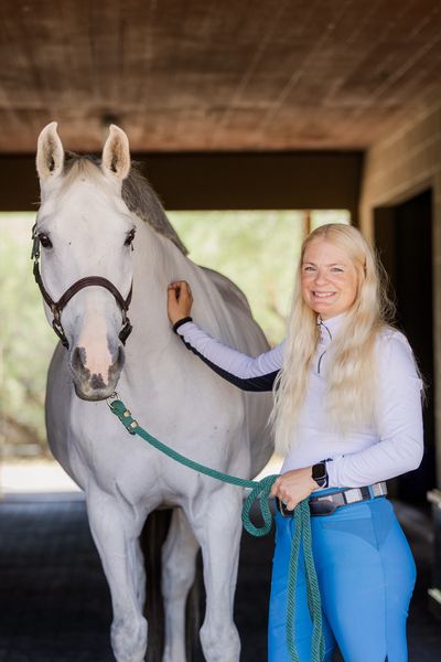 A smiling woman stands beside a white horse in a stable.