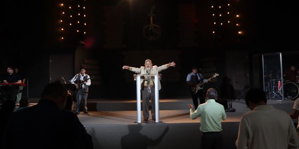 A preacher delivering a sermon on stage with musicians and attendees worshipping.