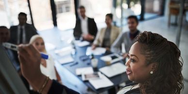 A woman leads a business meeting, writing on a glass board, with colleagues attentively listening.