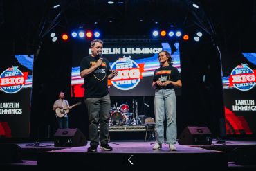 Two hosts on stage at a country music event with vibrant lights and screens.