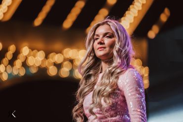Blonde woman with long hair in a sparkling pink outfit under warm lights.