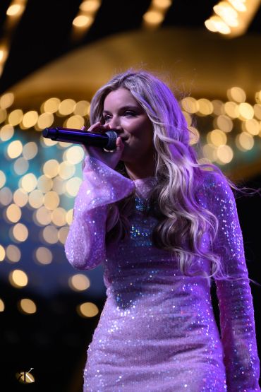 Female singer performing on stage with sparkling dress and colorful lights.