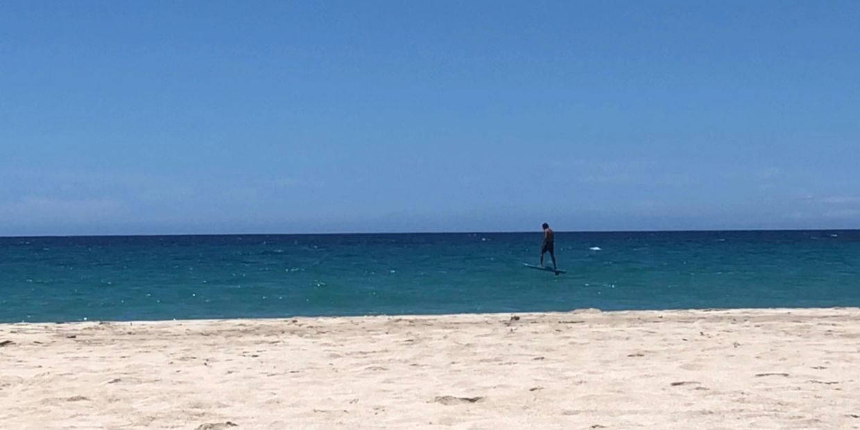 Person hydrofoiling on calm ocean near a sandy beach.